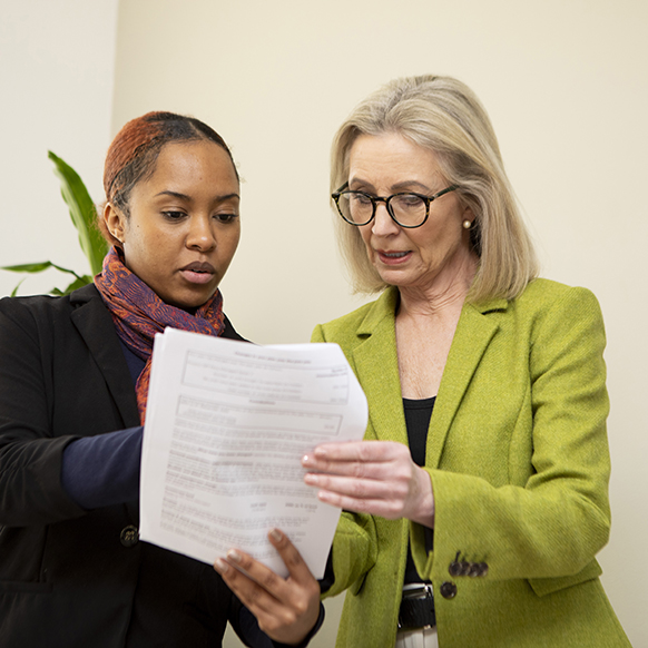 Two women in office hallway discussing something while looking at something on the paper they are holding