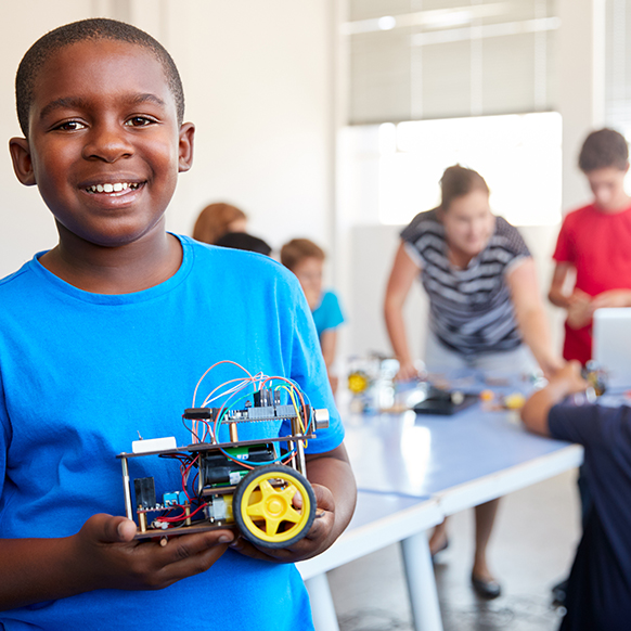 Classroom environment with a teacher in the background with young kids and one boy in front holding what he built