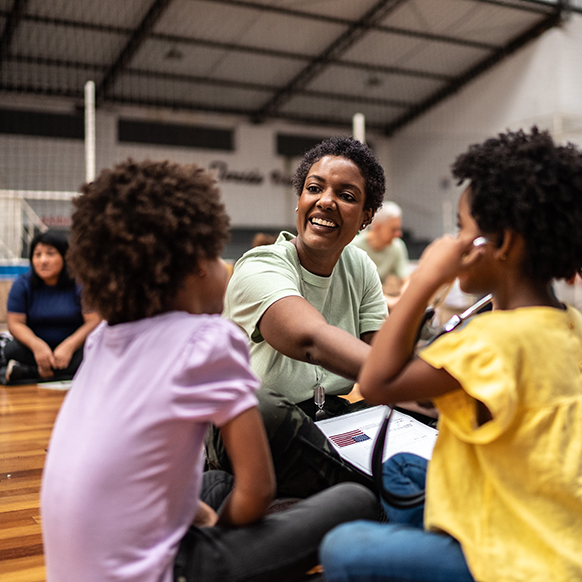 African American woman helper sitting on community center gym floor with 2 children talking