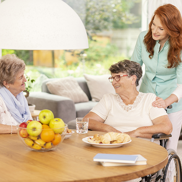 Senior ladies at table chatting as caregiver looks on to assist